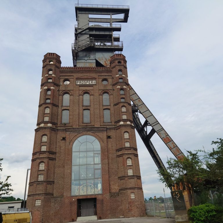 Historisches Fördergerüst (Malakoffturm) mit rotbrauner Backsteinfassade und modernem Aufzugsturm.